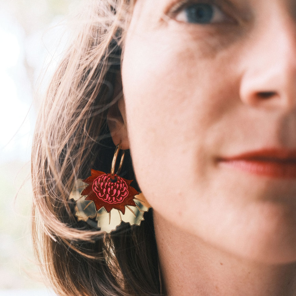 A close-up view of the midi waratah hoop earrings being worn. The frame is zoomed in close, so the gold and red earring is in focus while the woman wearing them is out of slightly focus.